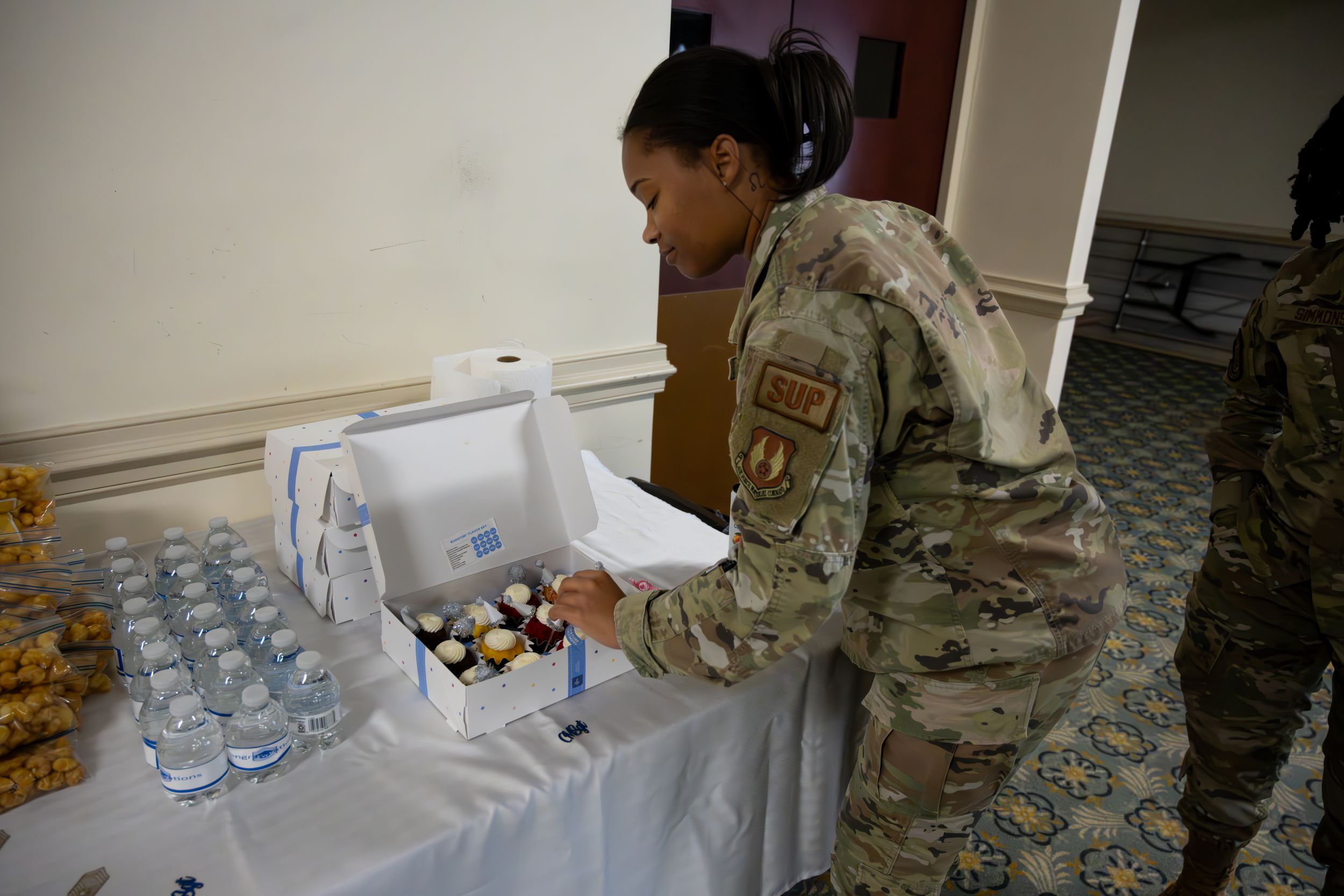 Military service members in uniform line up for refreshments at an indoor event.