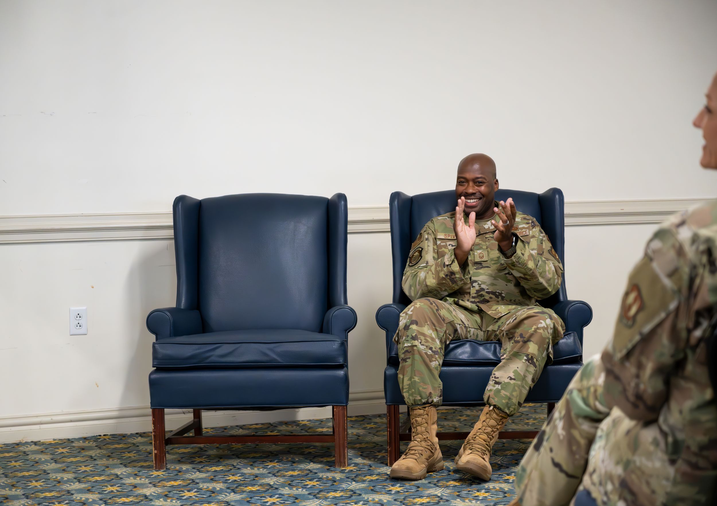Military personnel seated on blue chairs against a white wall during an indoor event.