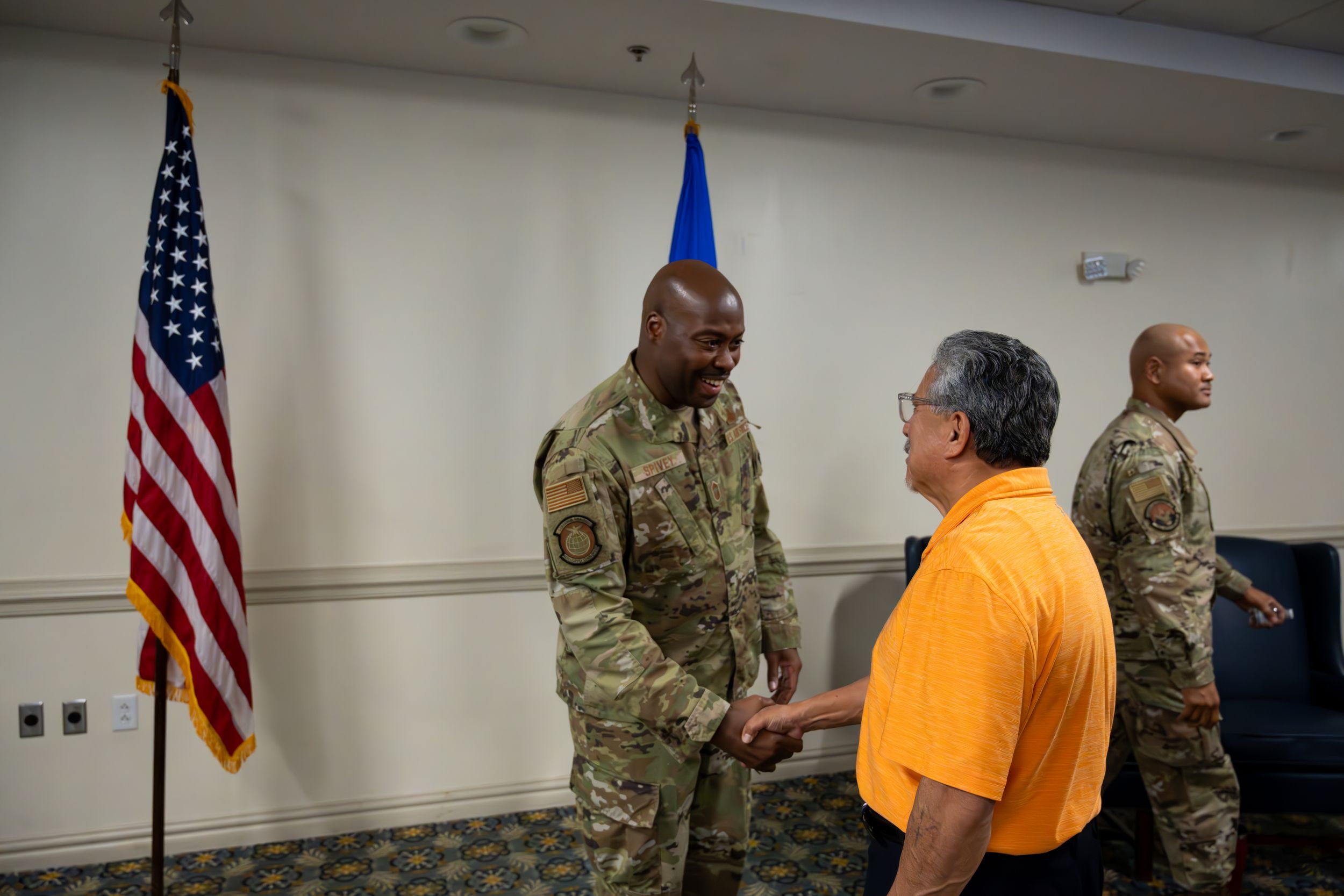 Military officer engaging in conversation with civilian in yellow shirt near American flag.