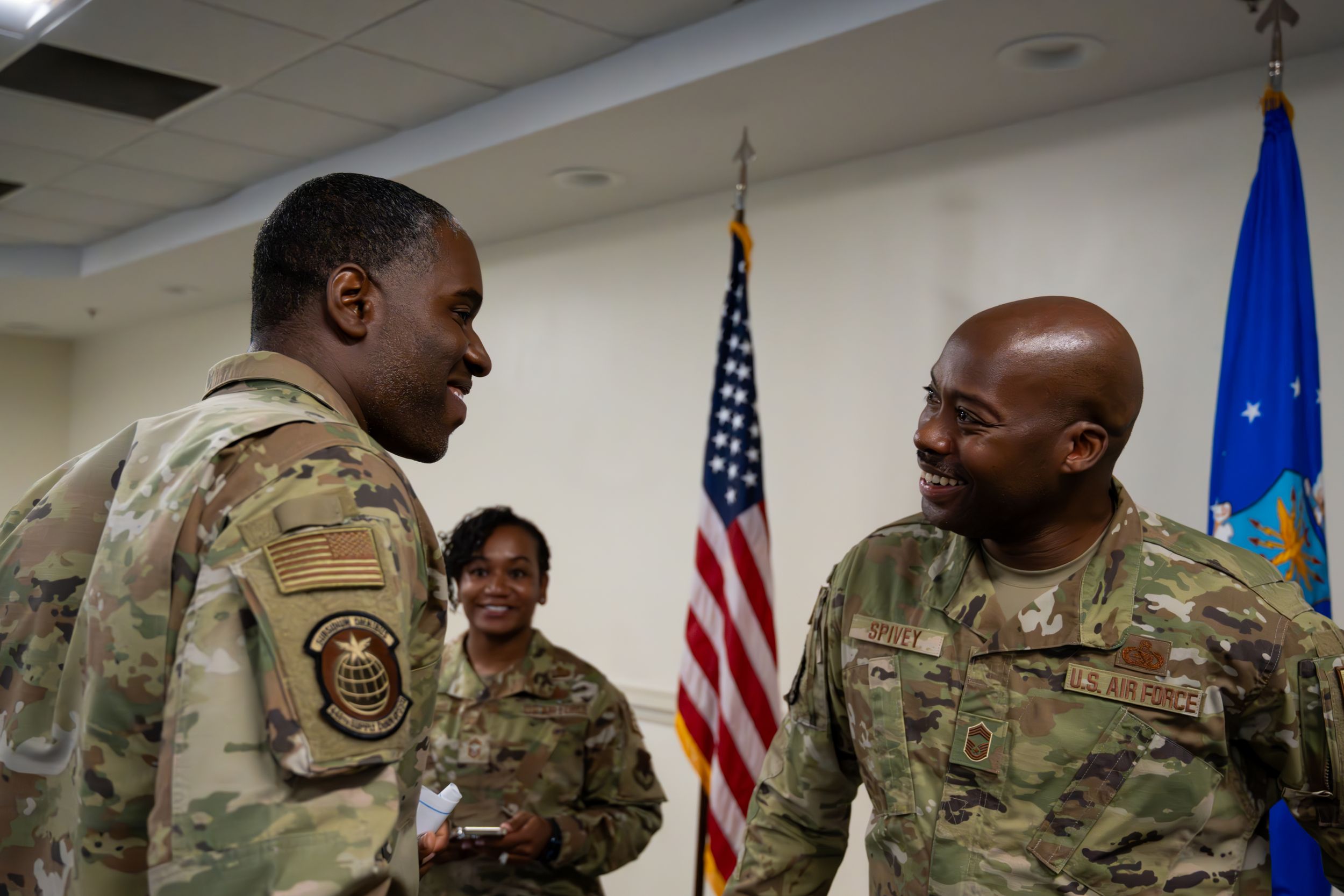 Service members engaged in discussion during military meeting with flags displayed.