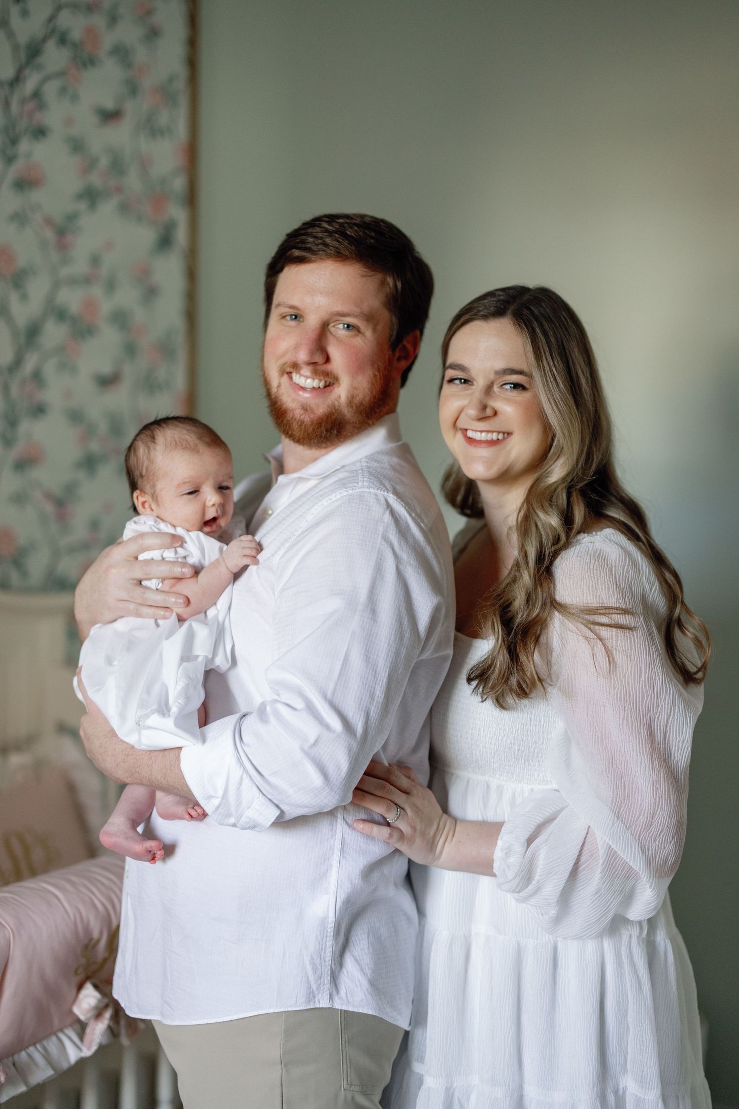 A family portrait in coordinating white outfits against a floral wallpaper backdrop.
