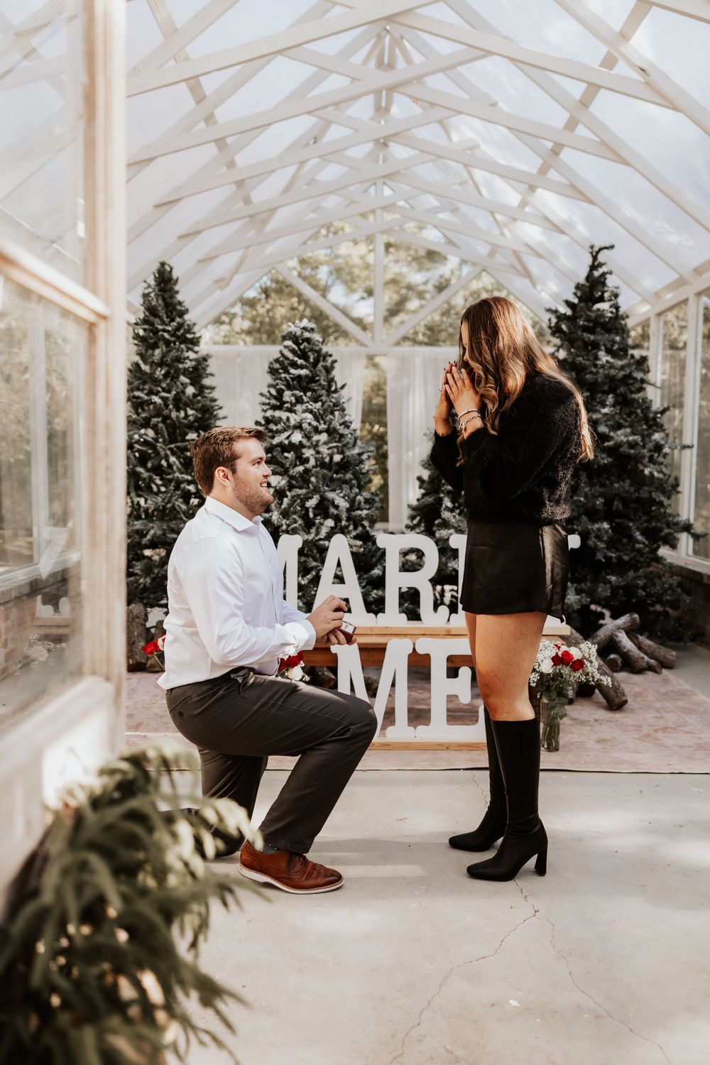 A wedding proposal scene in a greenhouse with Christmas trees and 'MARRY ME' letters.