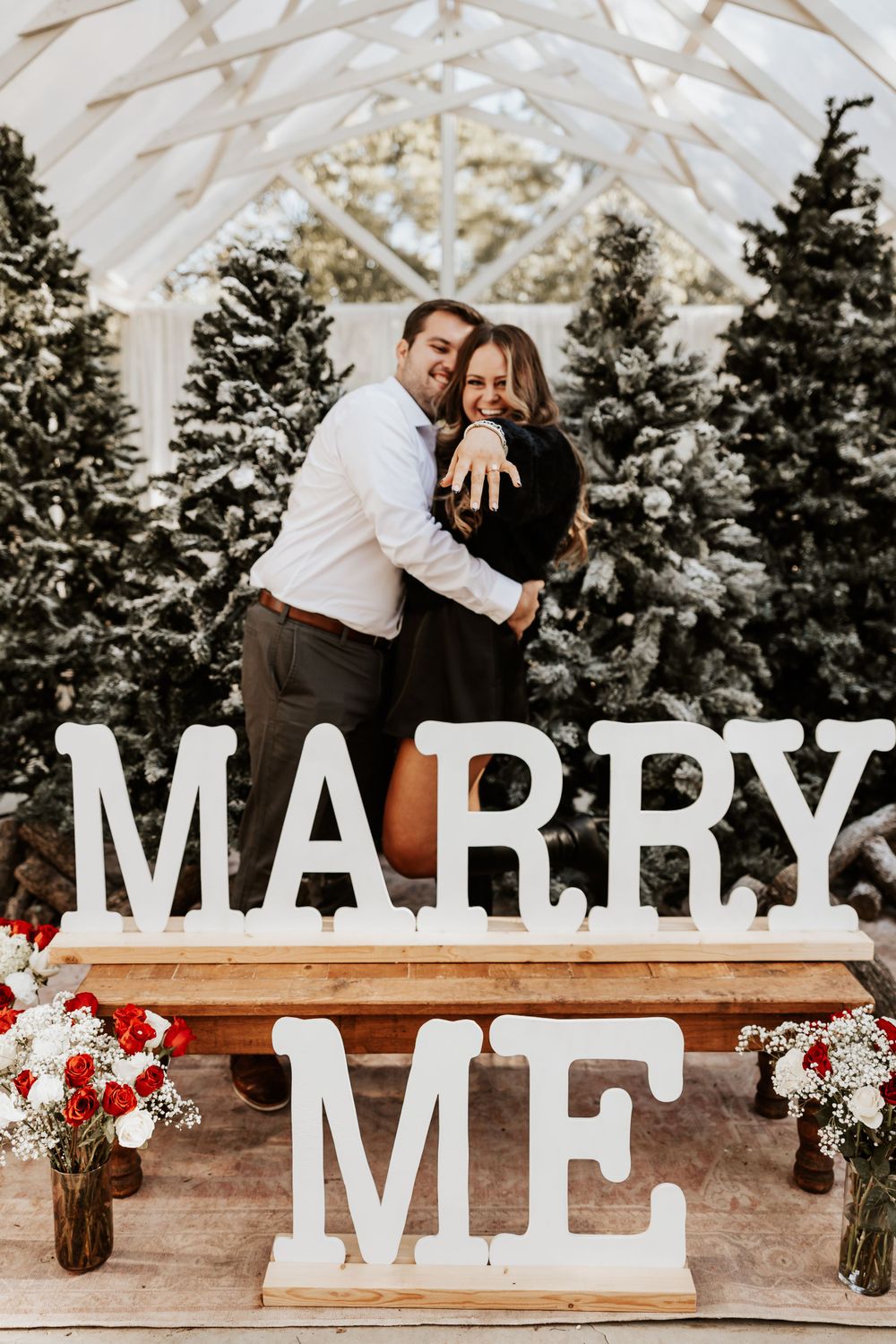 An engagement photo with a couple posing behind large 'MARRY ME' letters among Christmas trees.
