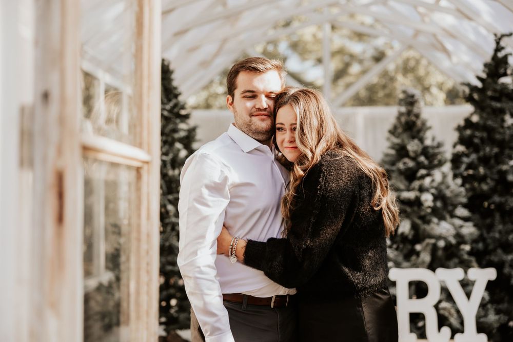A couple embraces in a greenhouse surrounded by decorated Christmas trees and white lights.