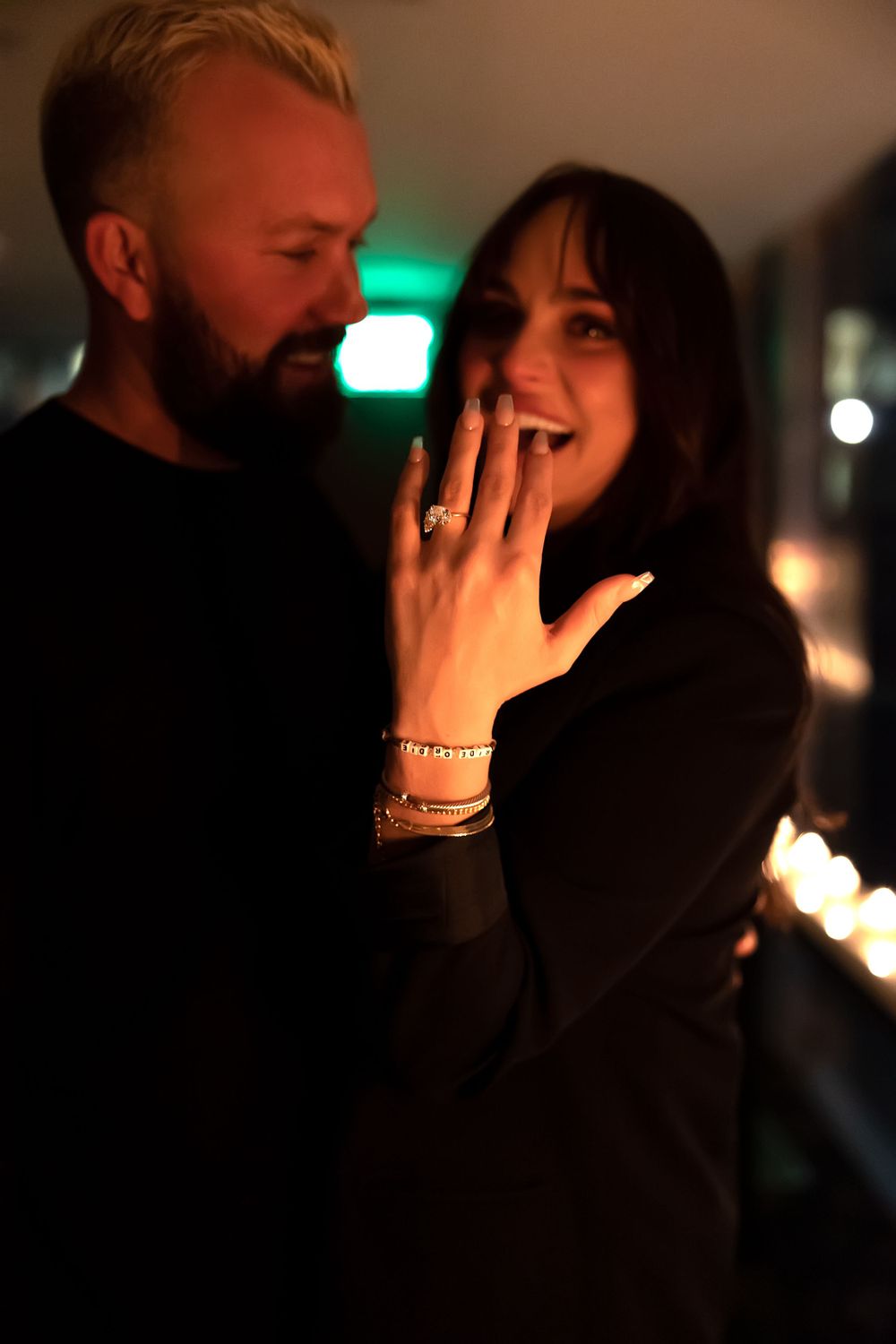 A dimly lit romantic proposal moment showing hands with jewelry and rings in soft lighting.