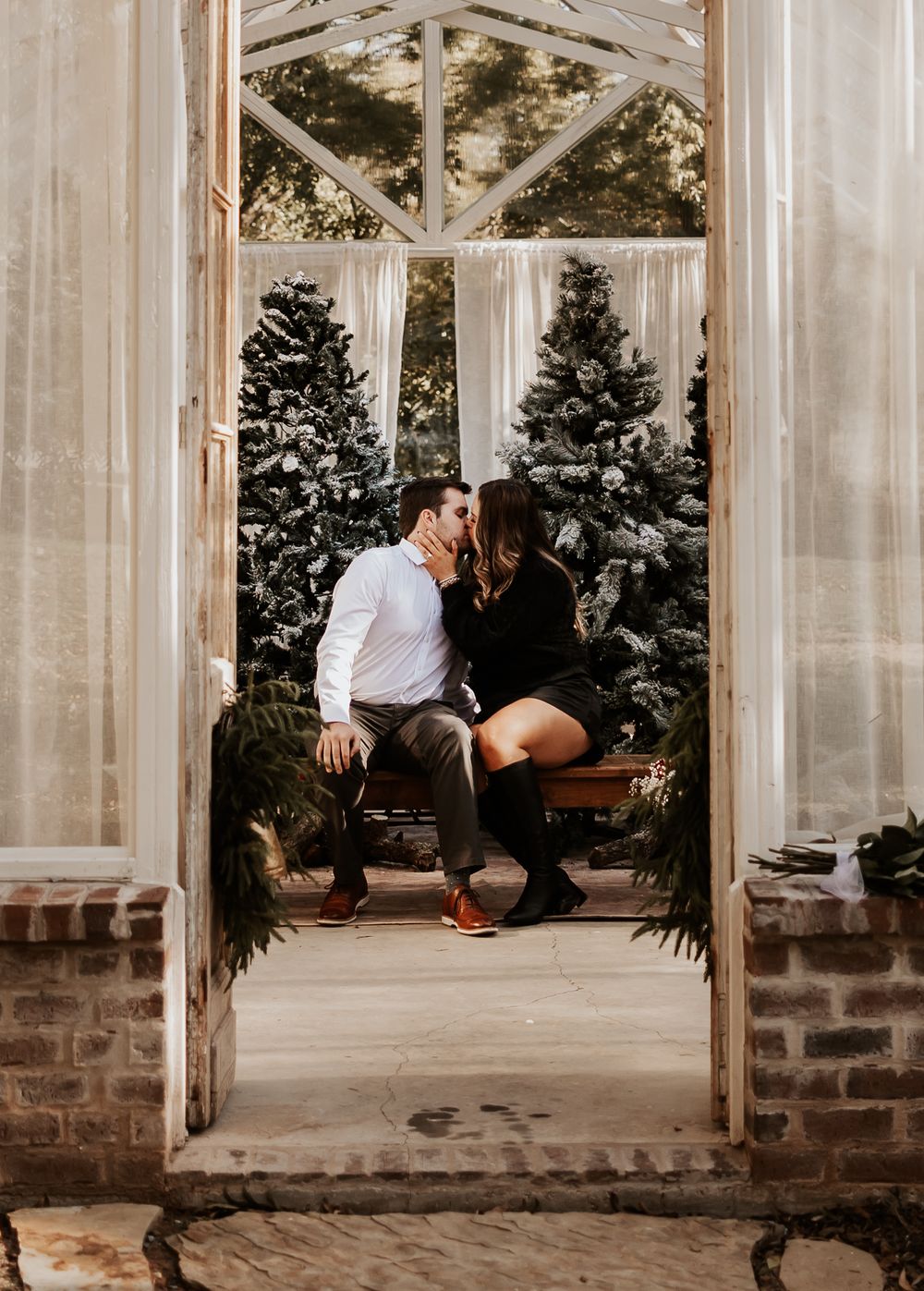 A couple shares an intimate moment in a greenhouse doorway framed by evergreen trees.