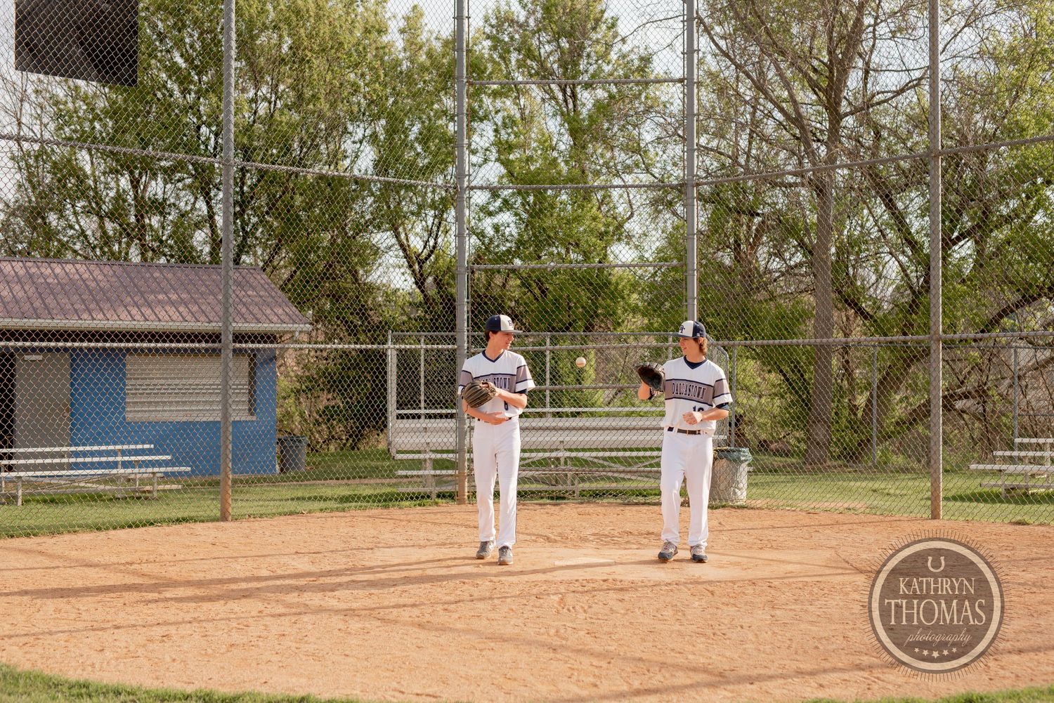 Baseball players standing on dirt field during game with buildings visible in background.