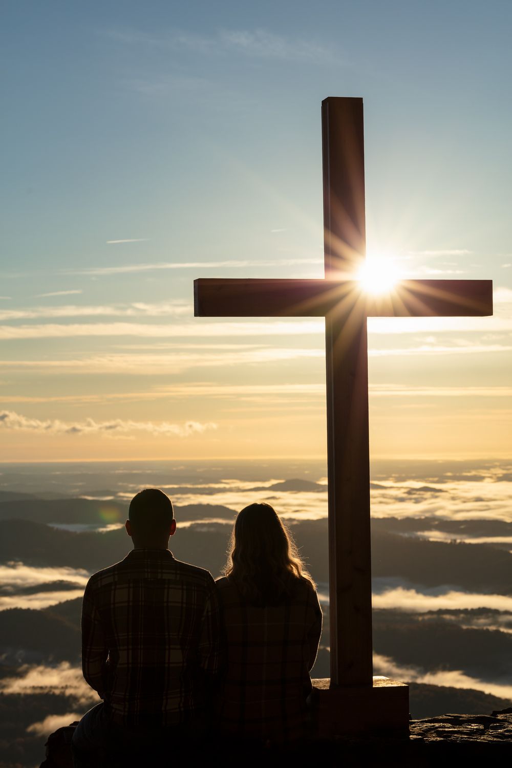 Silhouette of people near large crosses at sunset with clouds below mountain summit. Engagement photo at Pretty Place in South Carolina.