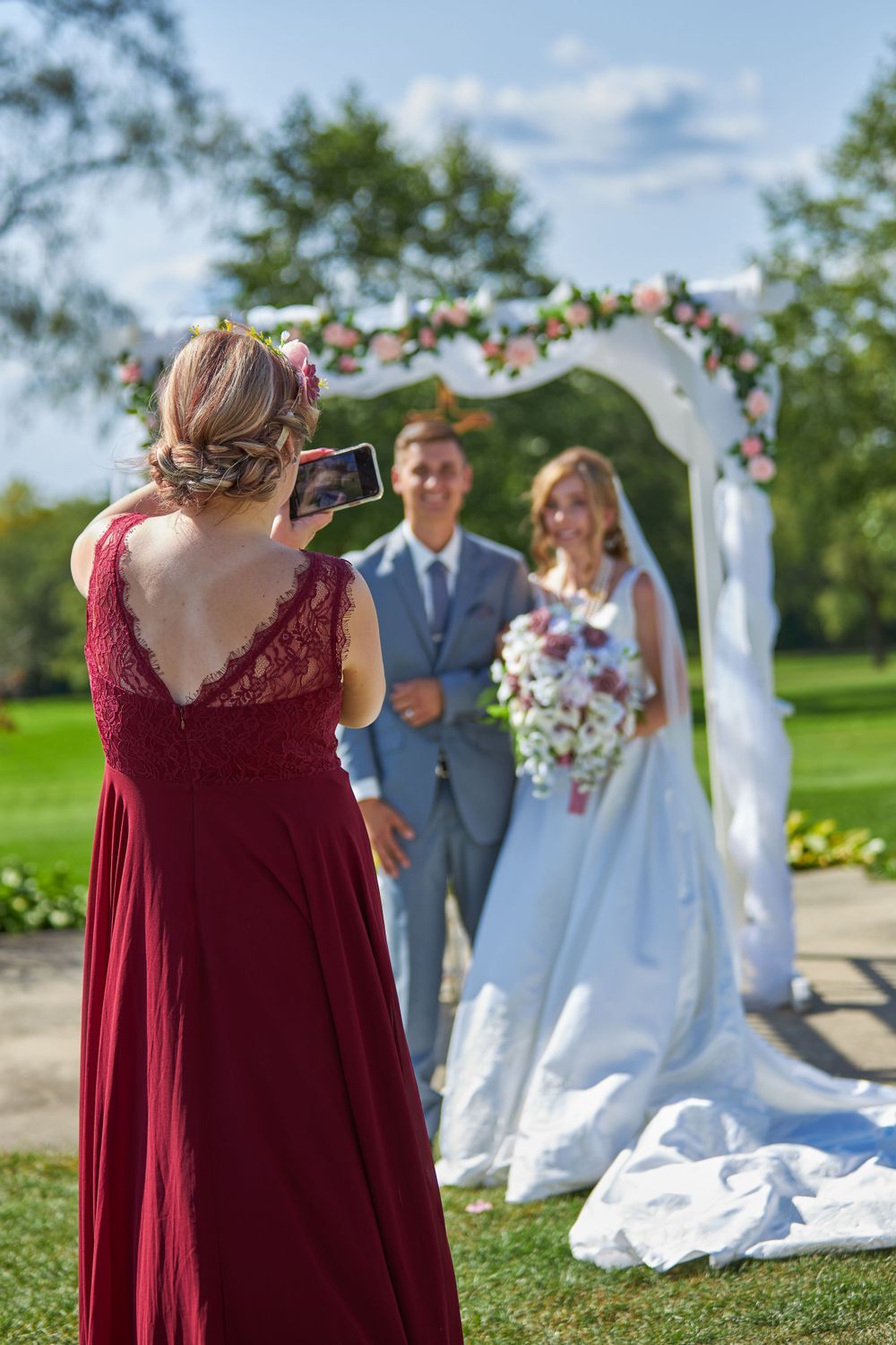 Wedding guest in burgundy dress taking photo of couple under floral arch.