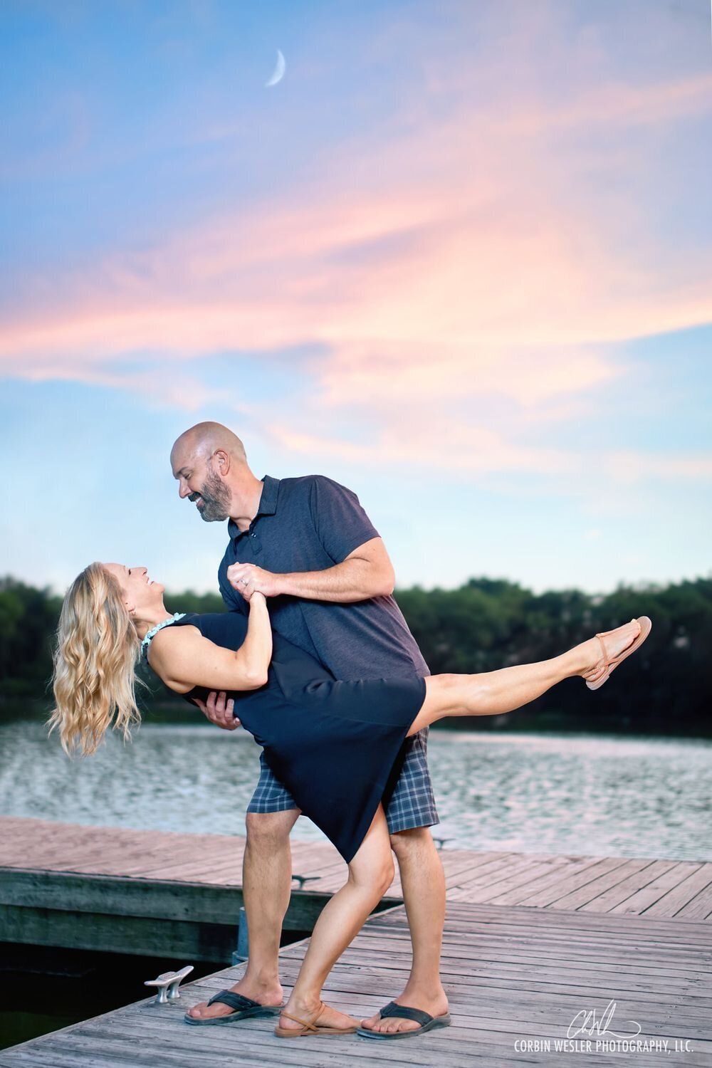 Couple performs romantic dance poses on wooden dock at sunset.
