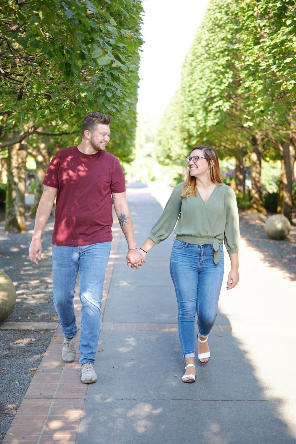 Couple in casual attire walking hand in hand down a tree-lined pathway on a sunny day.