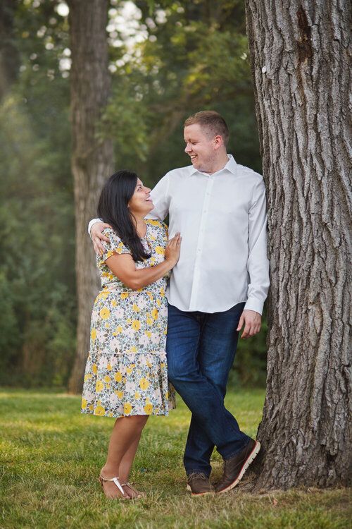 Couple in casual attire poses playfully among pine trees in natural setting.