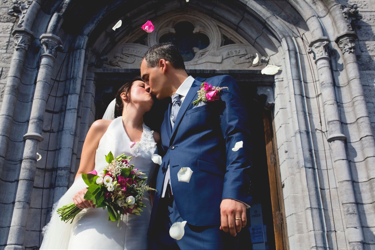 Newlyweds share intimate moment under gothic stone archway as rose petals fall.