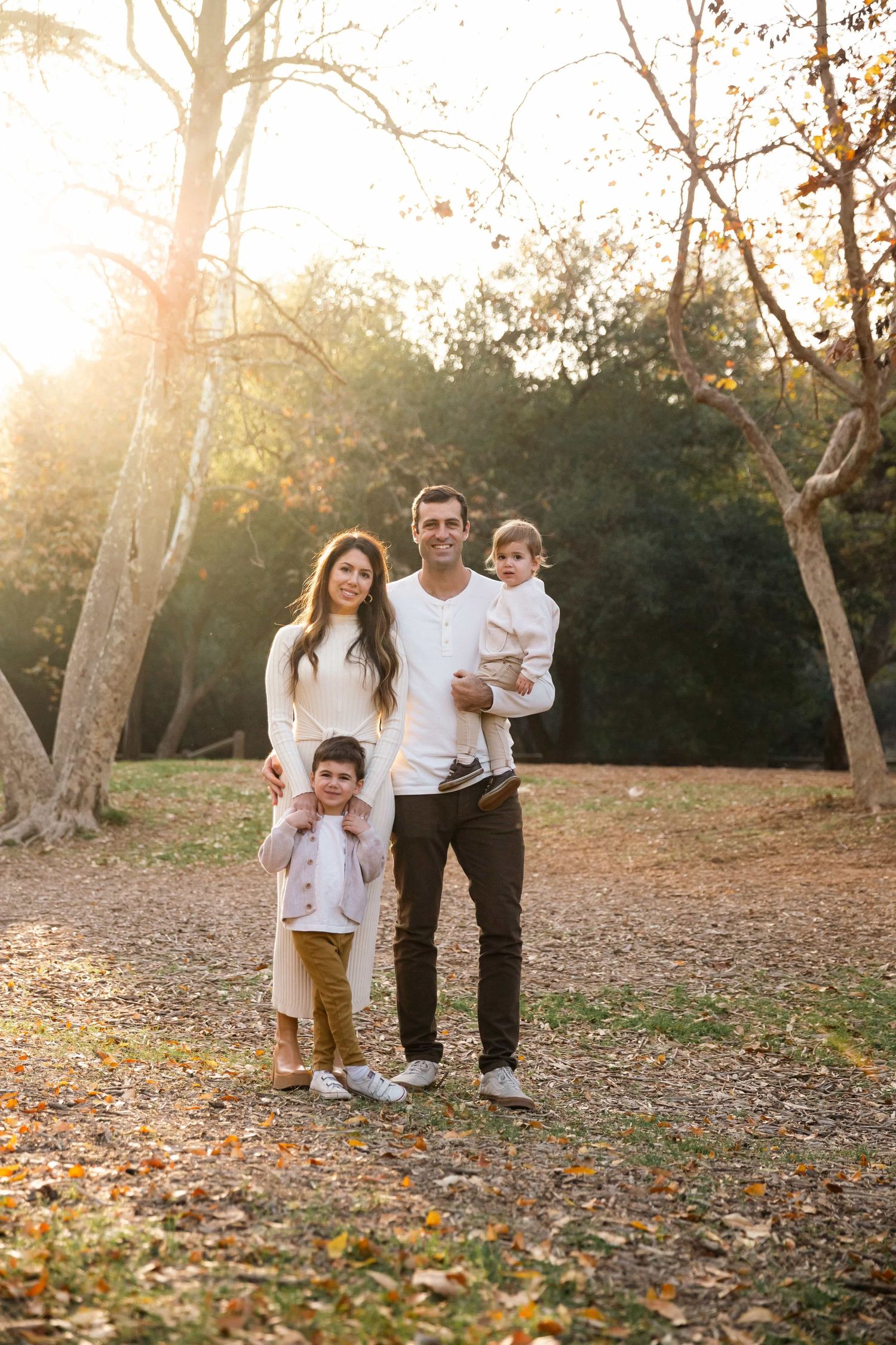Happy family of four posing in autumn park, parents with two young children in golden sunlight.