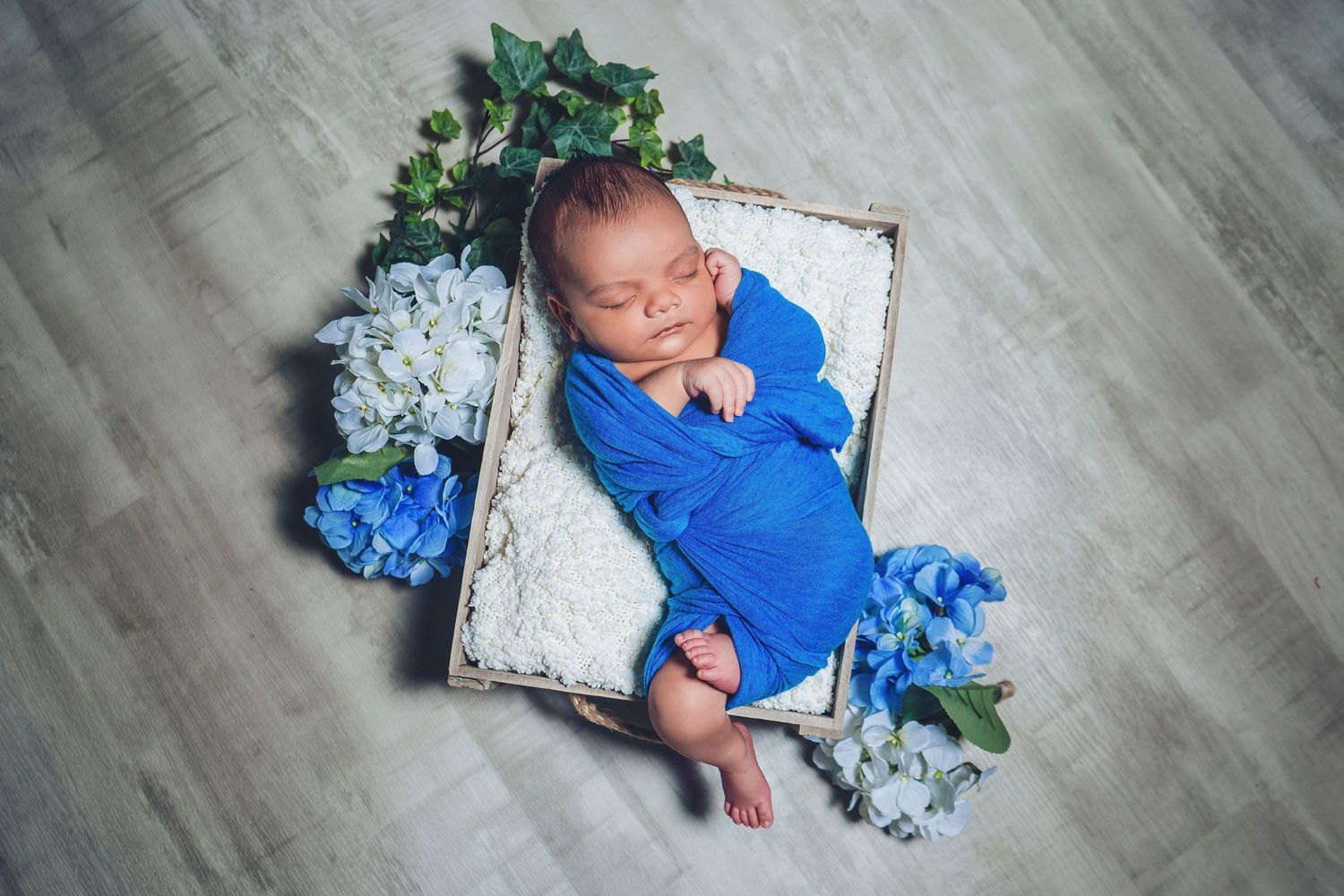 Sleeping newborn in blue wrap posed in white basket with blue hydrangea flowers.