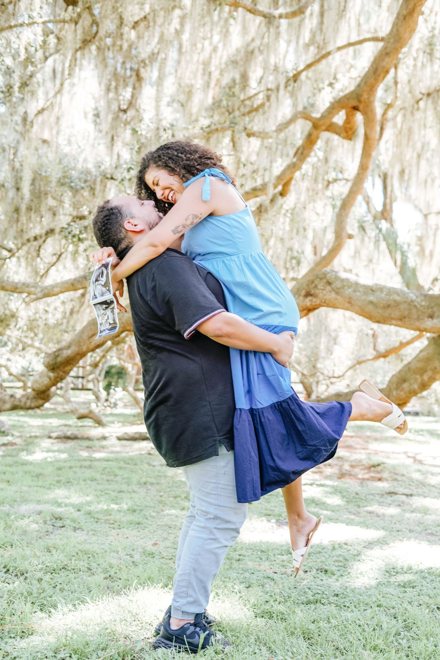 A romantic couple embraces joyfully under a large tree with hanging moss.
