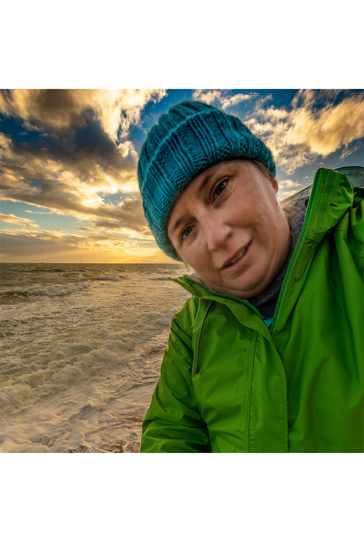 Person in green jacket and blue beanie takes beach selfie during golden sunset.
