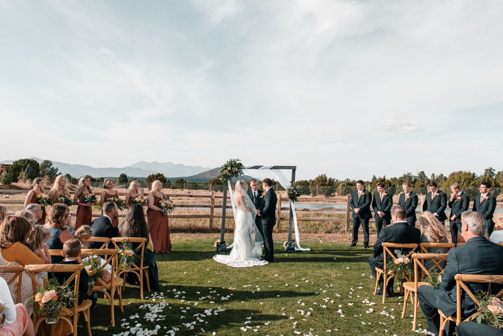 Rustic outdoor wedding ceremony with guests seated on wooden chairs against a mountain landscape.