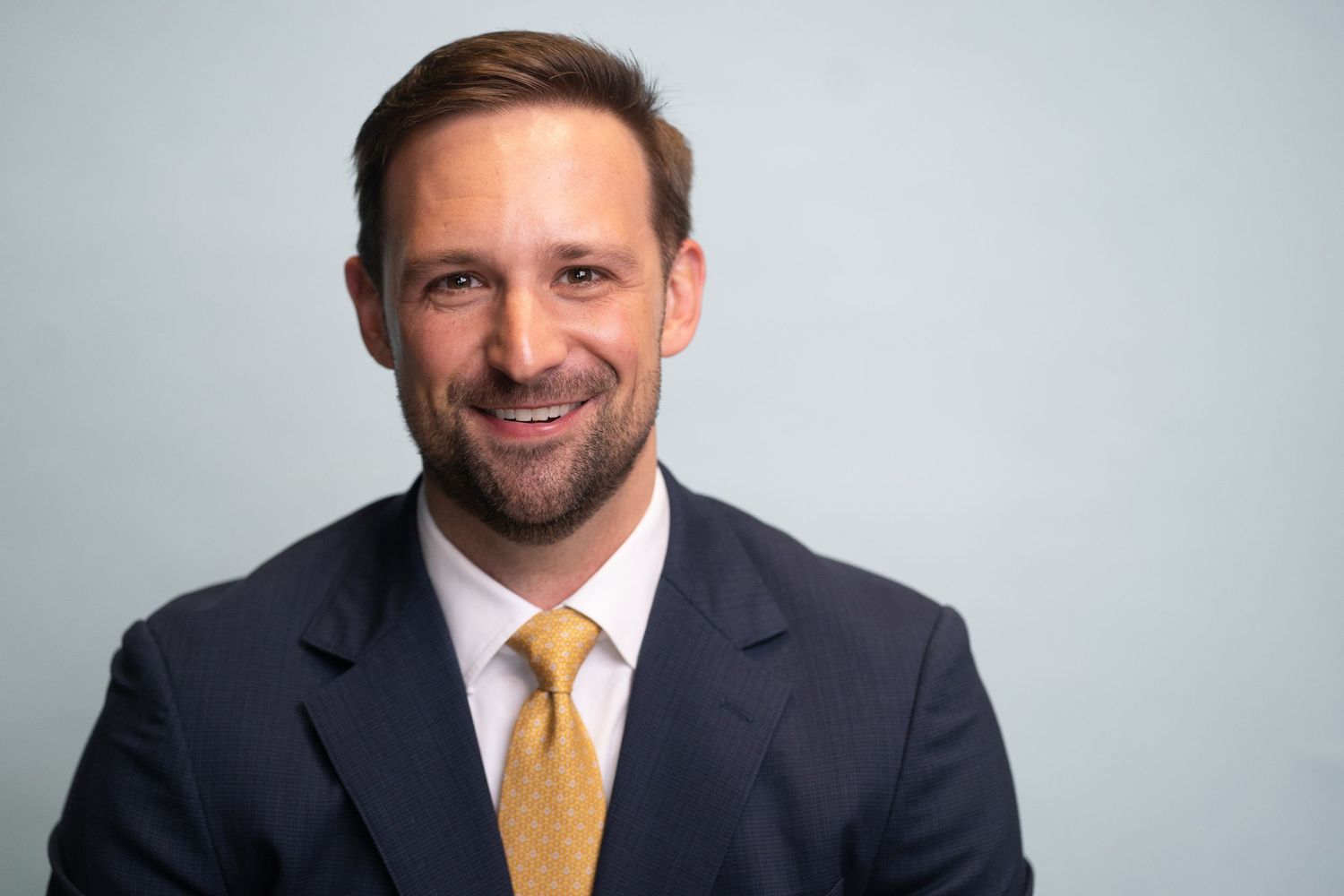 Professional headshot of businessman wearing dark suit and yellow tie against light background.