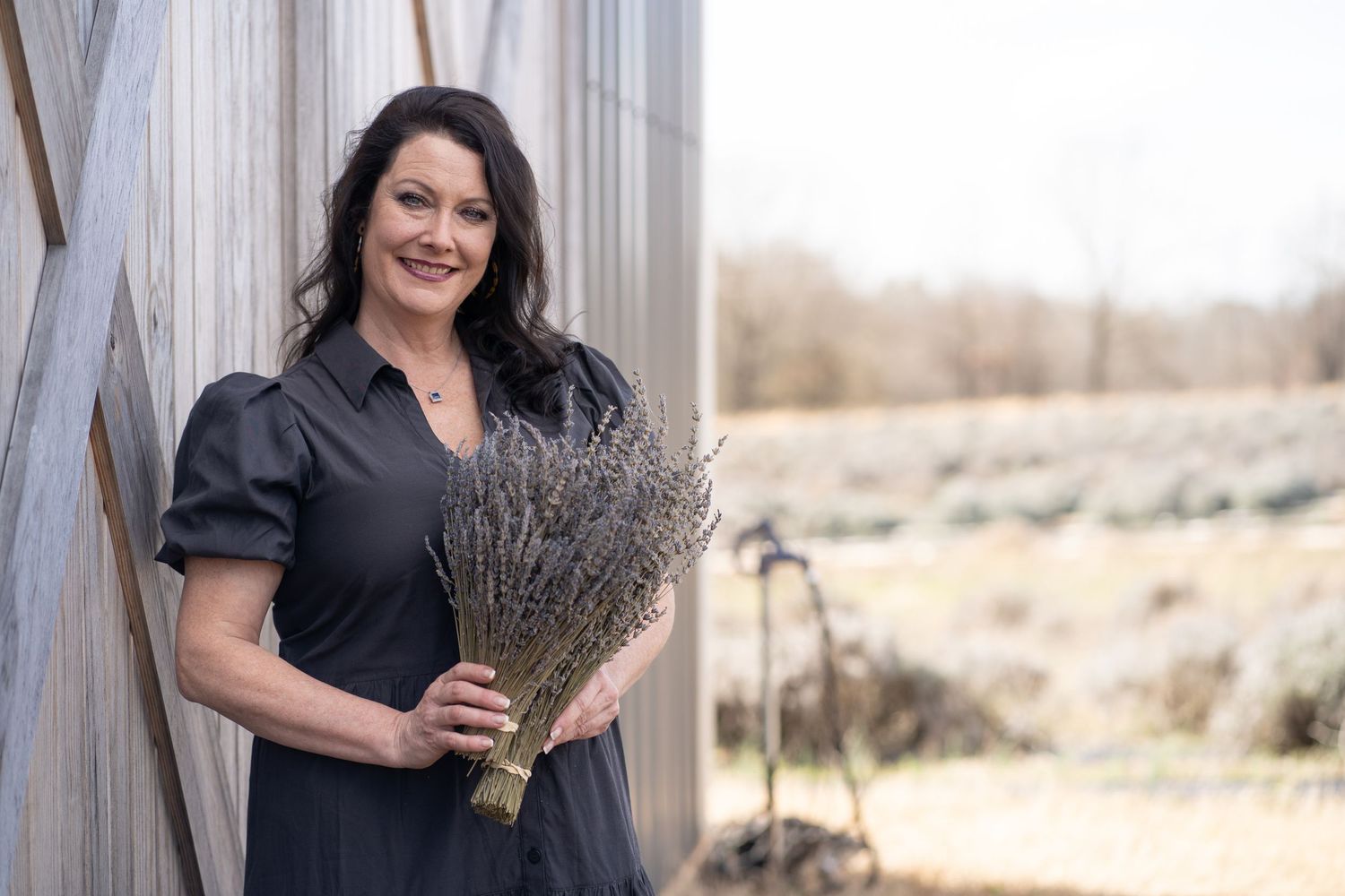 Woman in black dress holds dried lavender bouquet while standing against rustic backdrop.