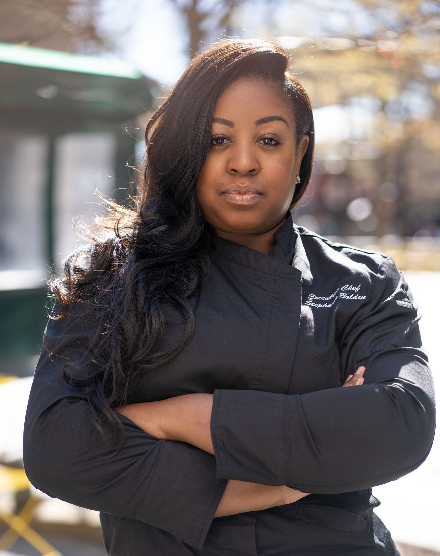 Professional in black winter coat poses confidently with arms crossed outdoors.