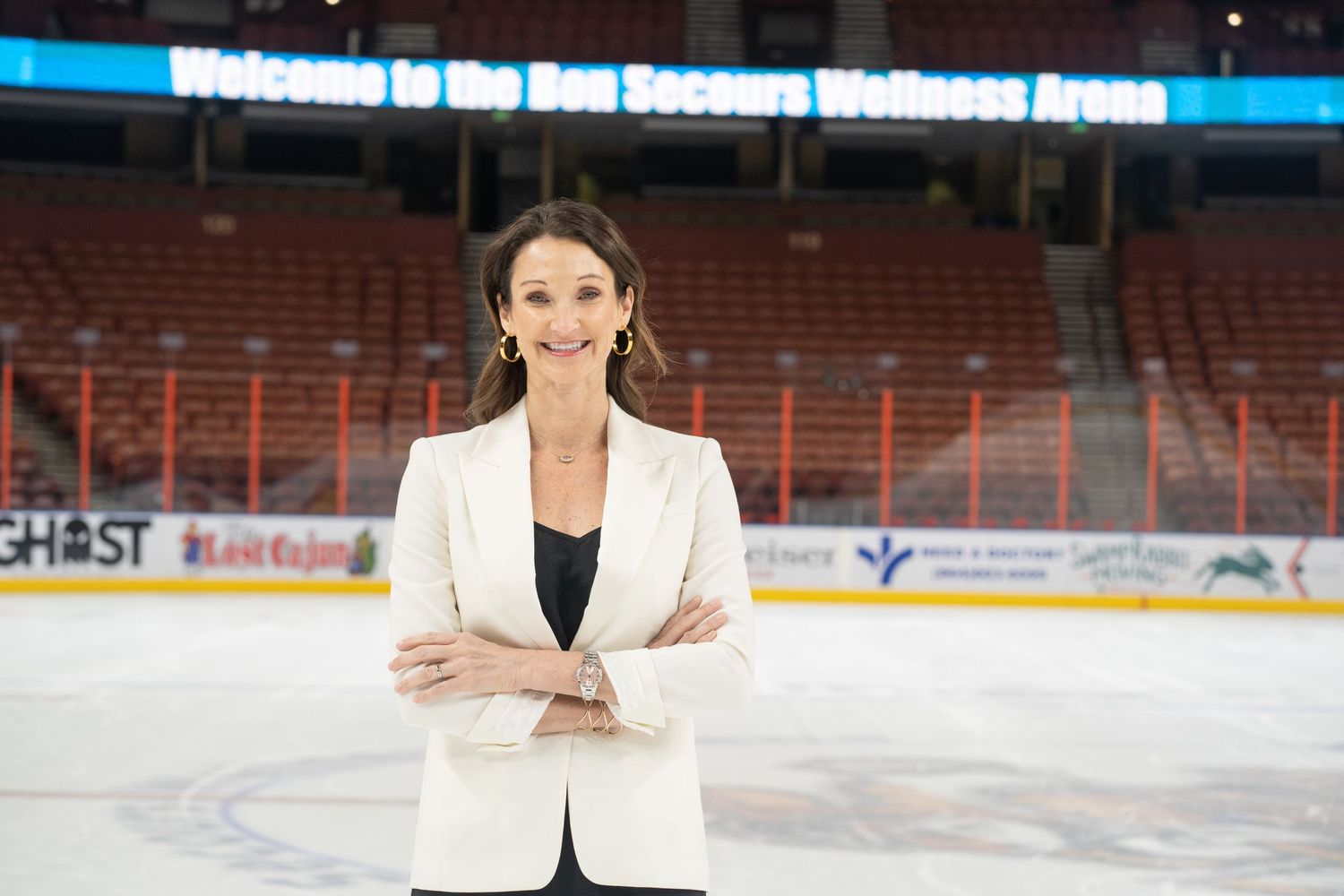 Professional woman in white blazer standing at ice rink with stadium seating in background.