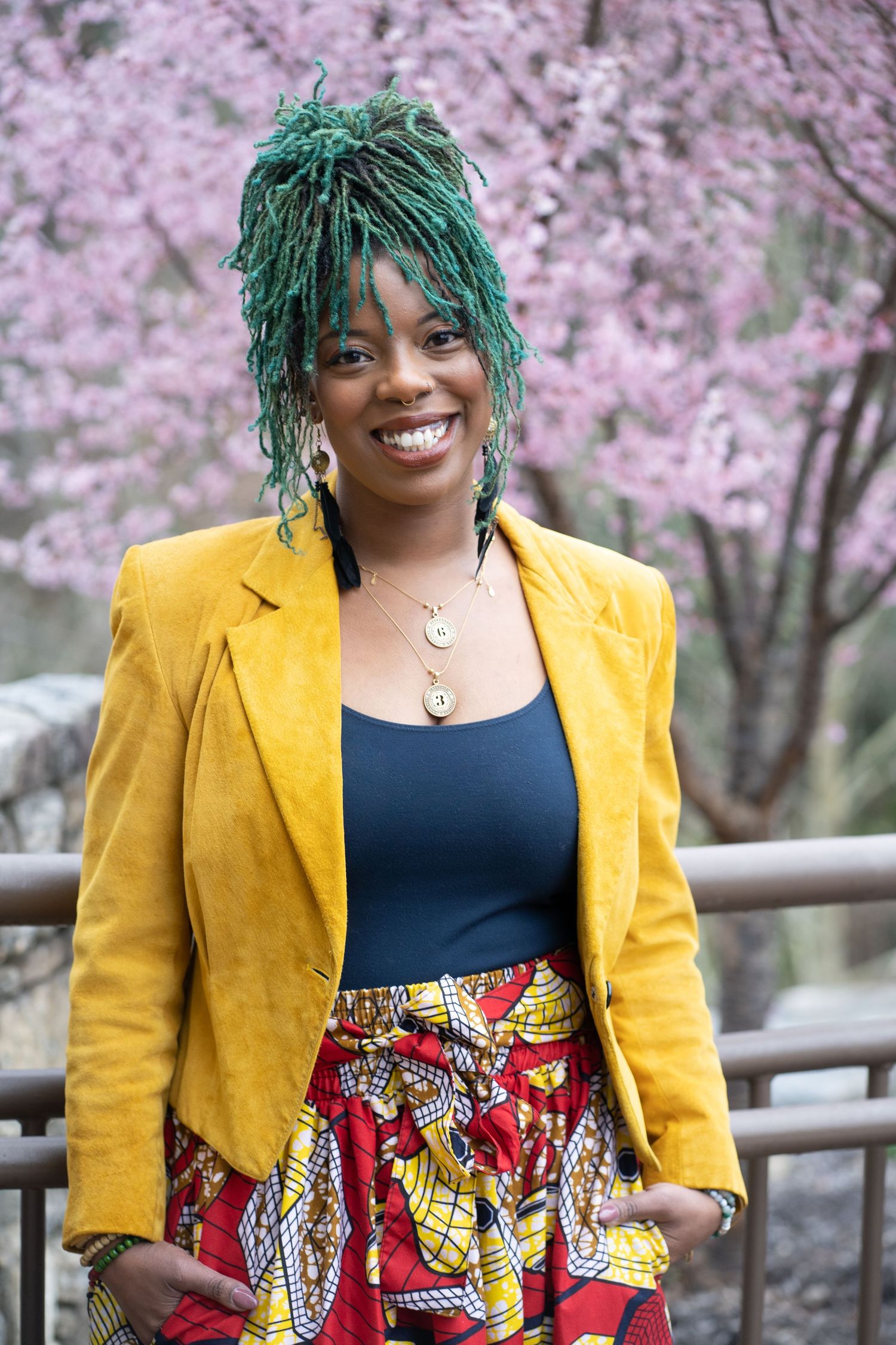 Vibrant portrait with green braids, yellow blazer, and patterned skirt against pink cherry blossoms in background.