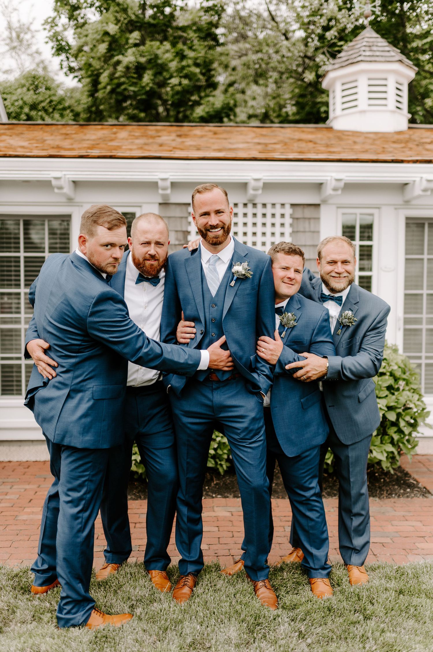 Groomsmen in matching navy suits pose playfully outside colonial building.