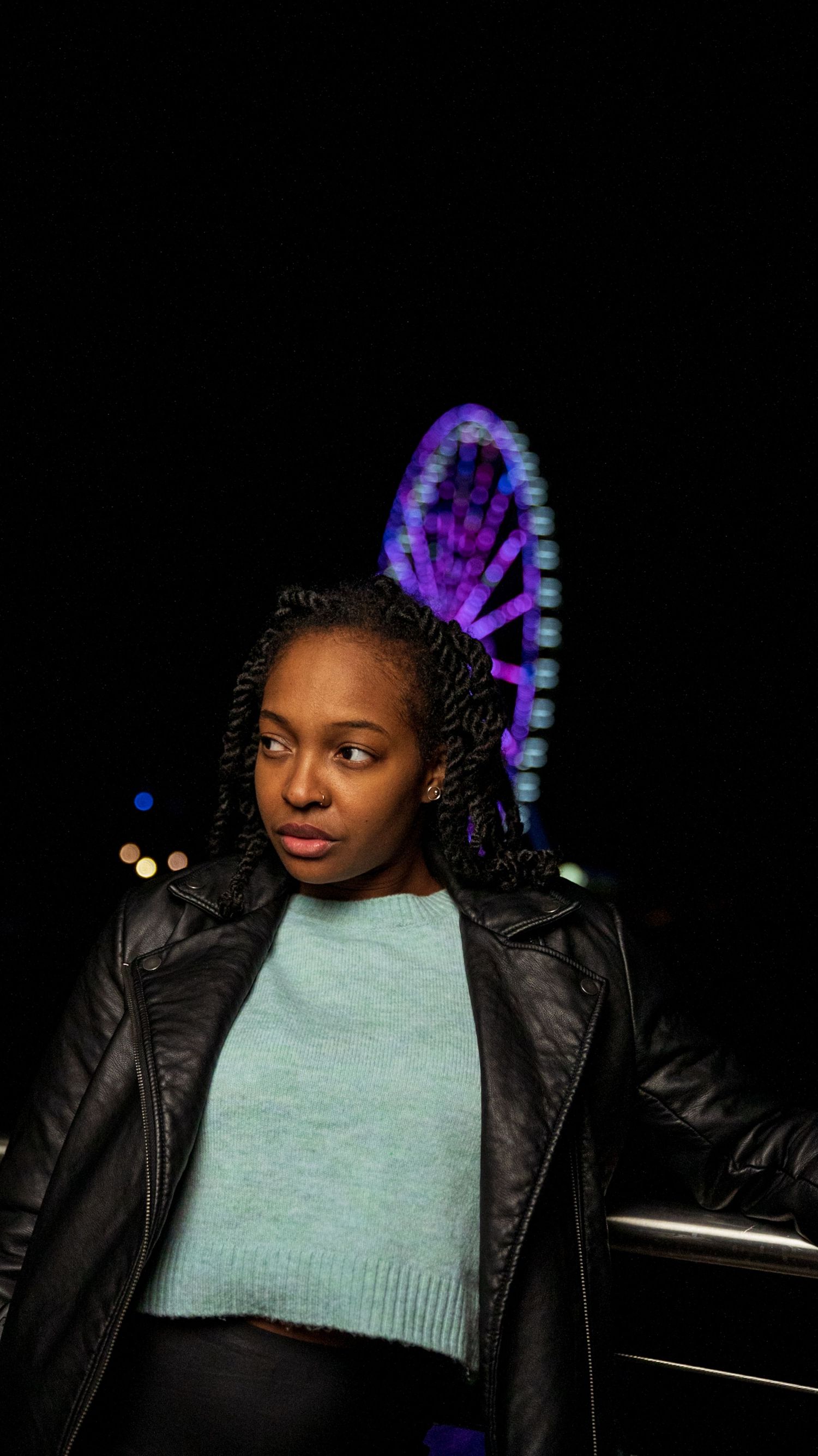 Young Black woman with braids wearing leather jacket and mint sweater posing at night near illuminated Ferris wheel.
