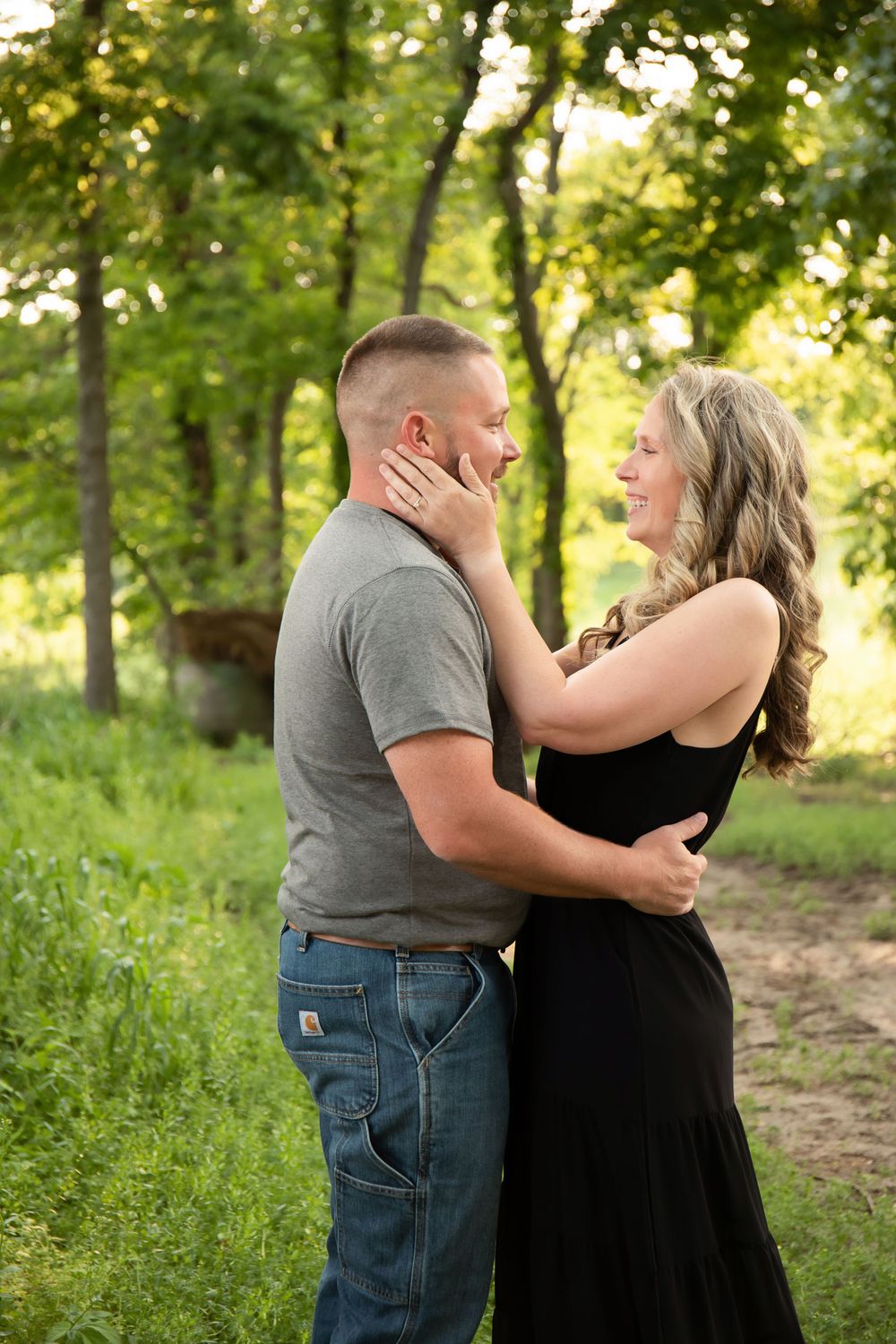 Intimate couple embraces tenderly in a sunlit wooded area with lush greenery.