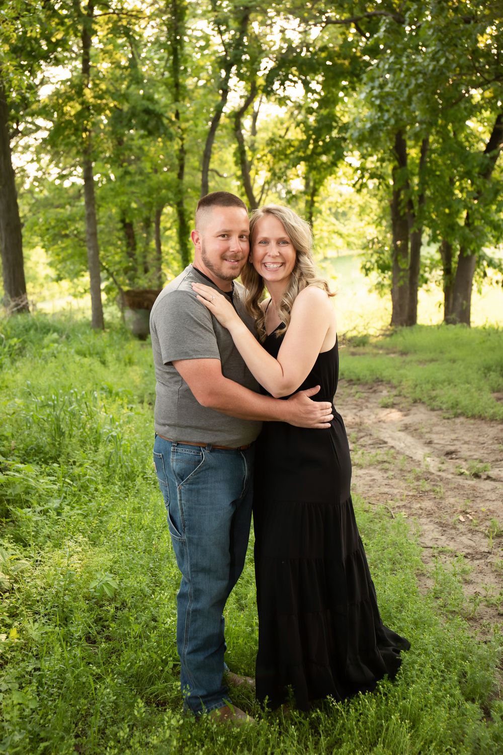 Happy couple embraces while standing on a dirt path surrounded by green trees and grass.