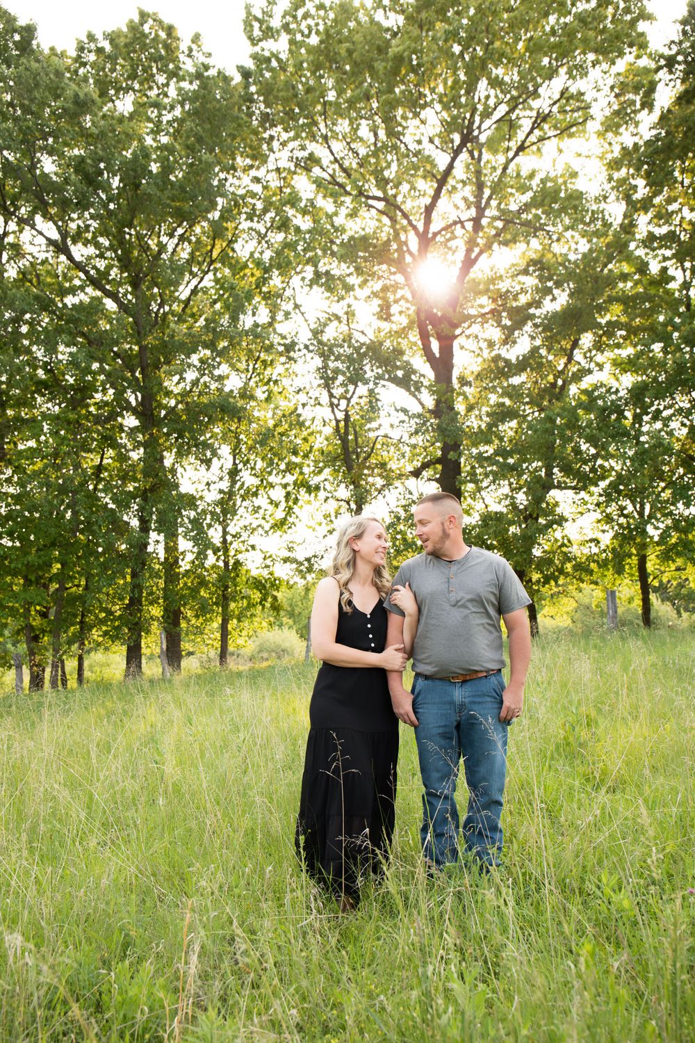Couple stands together in tall grass with sunlight streaming through trees behind them.