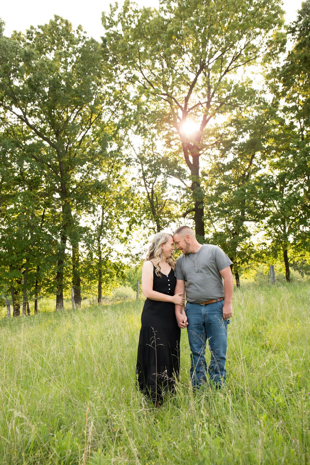 Couple shares an intimate moment standing in tall grass with sunlit trees creating a romantic backdrop.