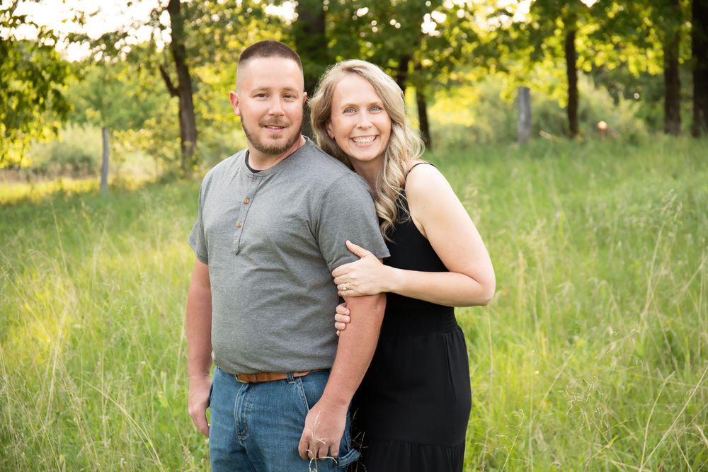 Couple poses casually together in a sun-drenched field surrounded by trees.