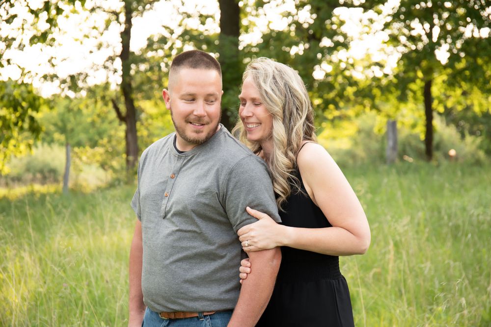 Couple laughs together while posing in a sunny meadow with trees in the background.