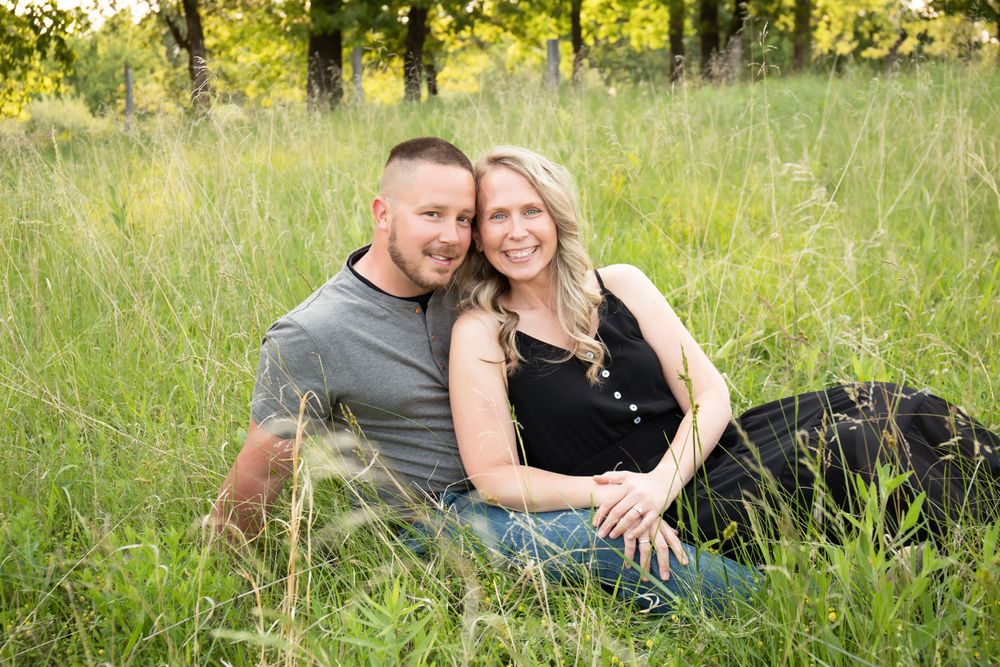 A couple sits together in a sunlit meadow with tall grass and trees in the background.