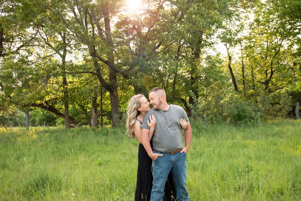 A couple stands together in a green field with sunlight streaming through trees.