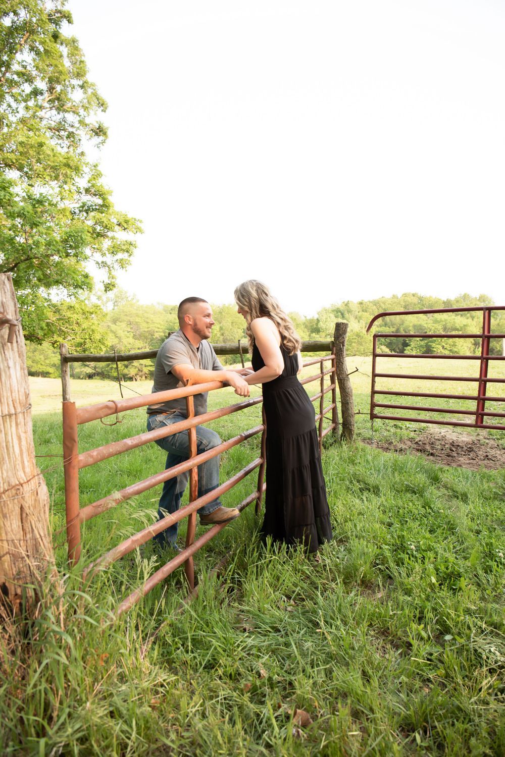 A couple shares a romantic moment by a rustic fence gate in a pastoral setting.