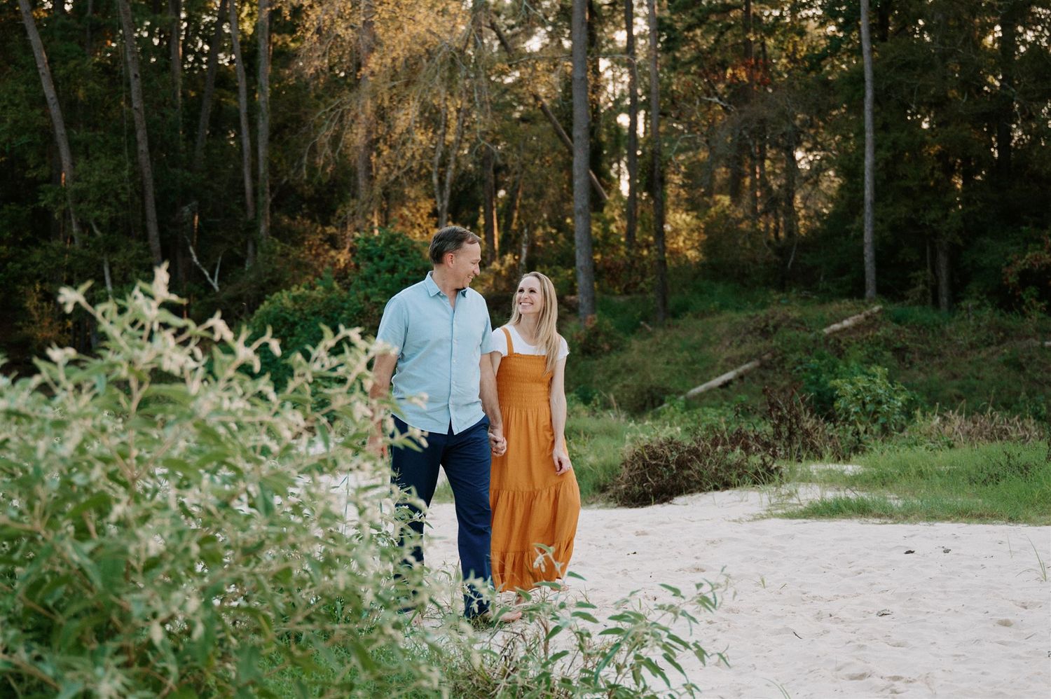A couple strolls along a sandy path surrounded by lush greenery and trees at golden hour.
