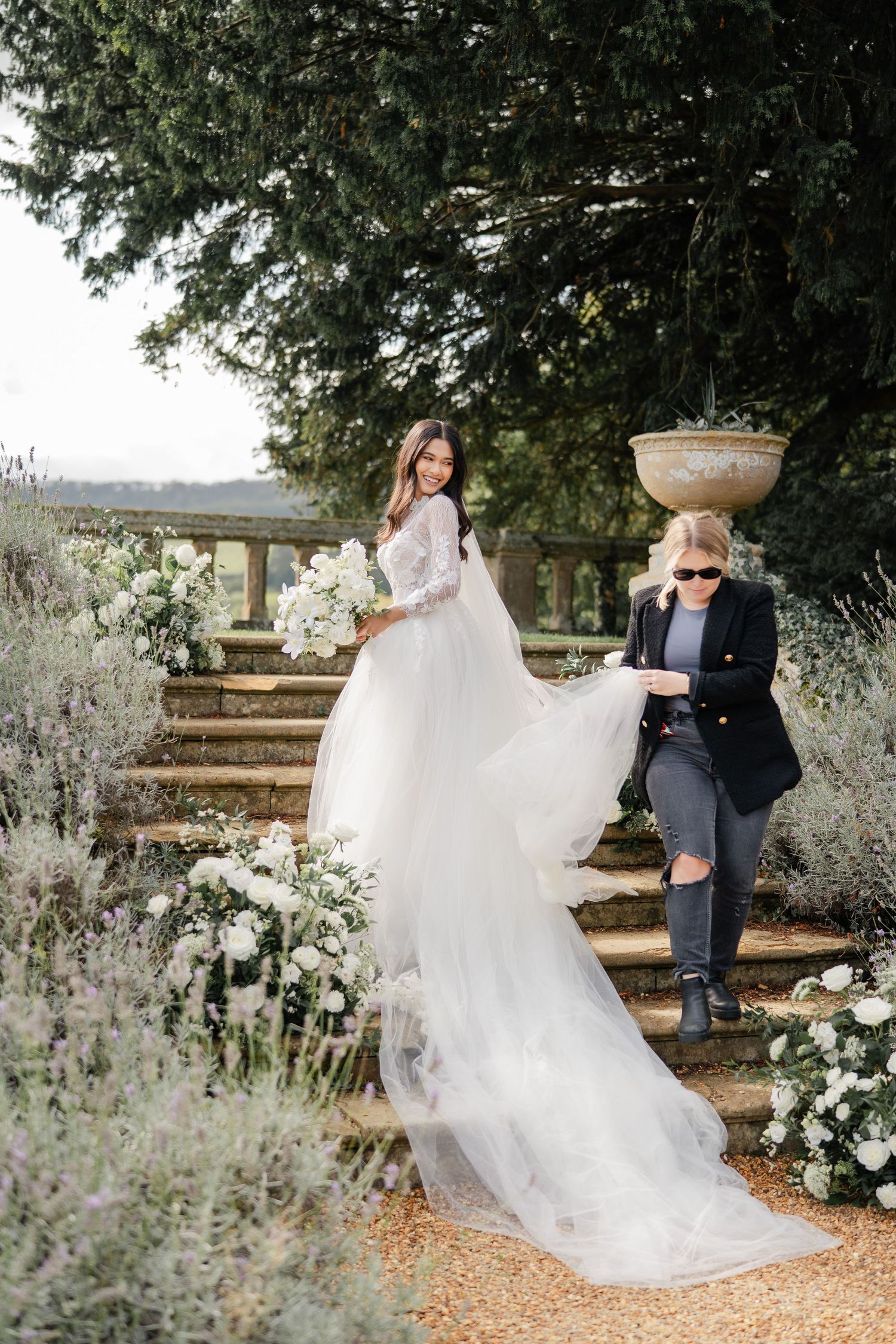 A couple poses on rustic garden steps surrounded by white floral arrangements.