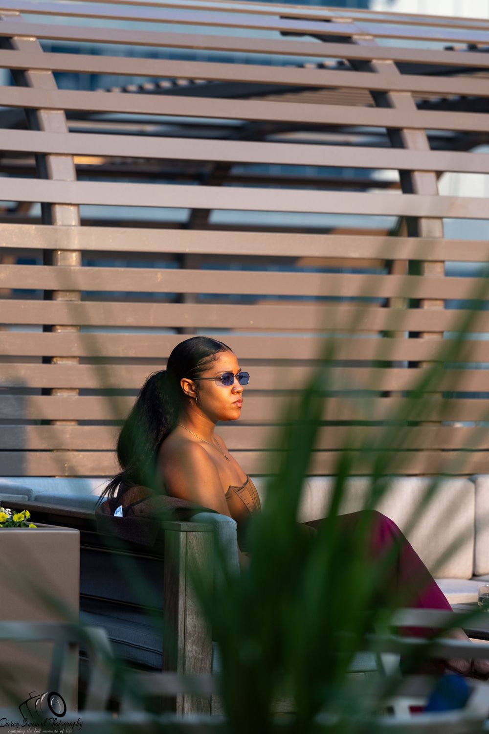 Person wearing sunglasses sits in bleachers on a sunny day with horizontal slats creating dramatic shadows and lines.
