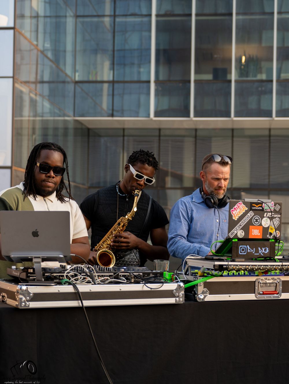 Three DJs perform at an outdoor event with laptops, turntables and a saxophone against a modern glass building backdrop.