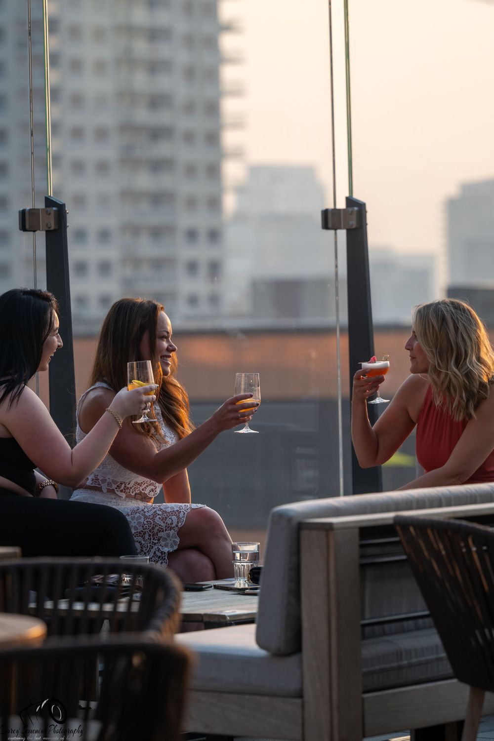 Group of friends enjoying drinks together on a rooftop patio at sunset with city buildings in background.