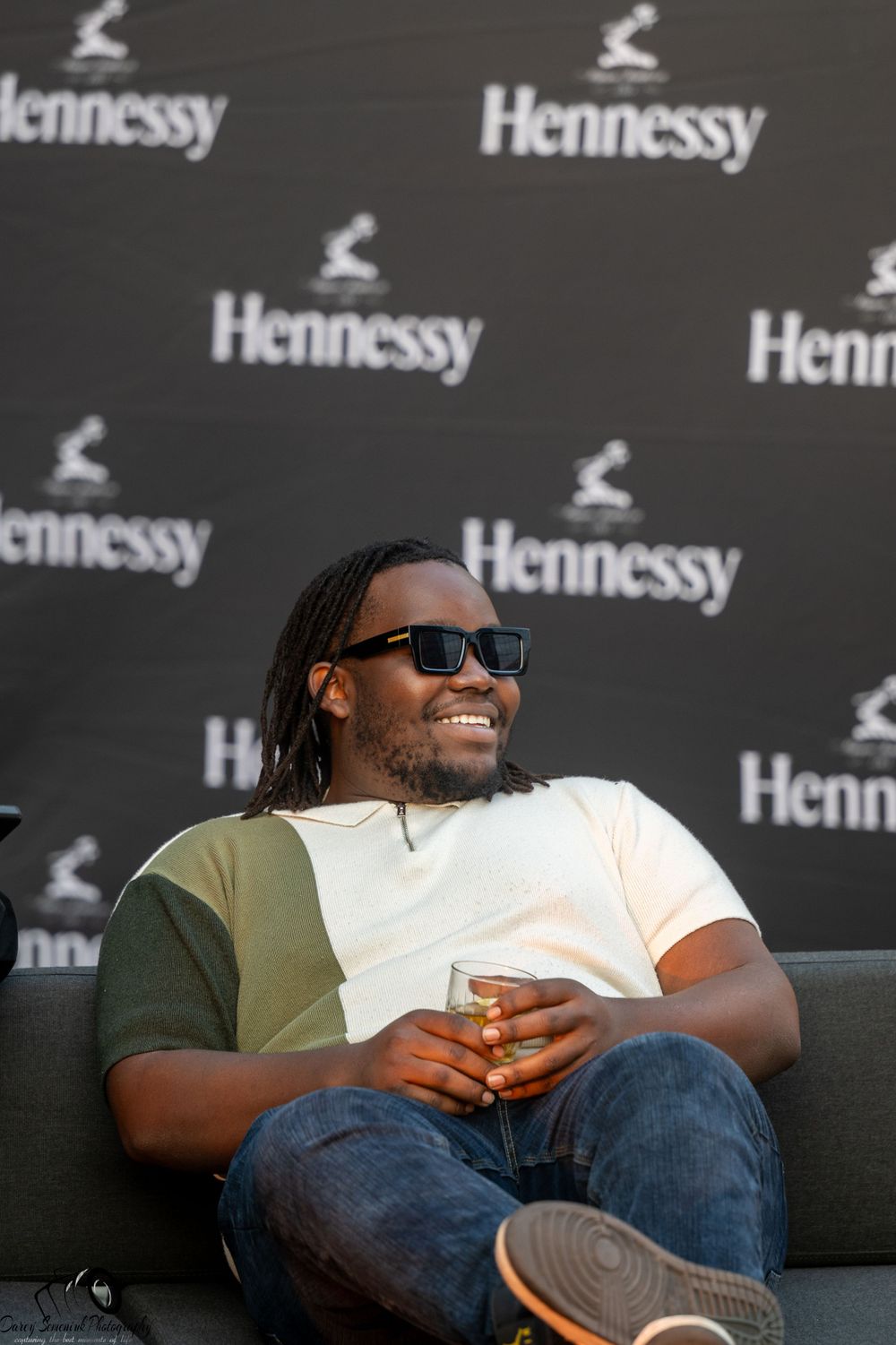 Person in sunglasses and casual attire laughing while sitting on a couch at a Hennessy-branded event.