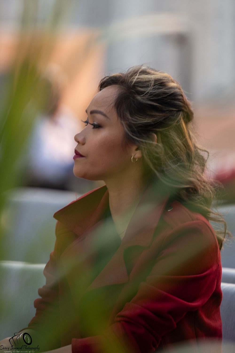 Person in red jacket sits outdoors with wind-blown hair against blurred natural background at sunset.