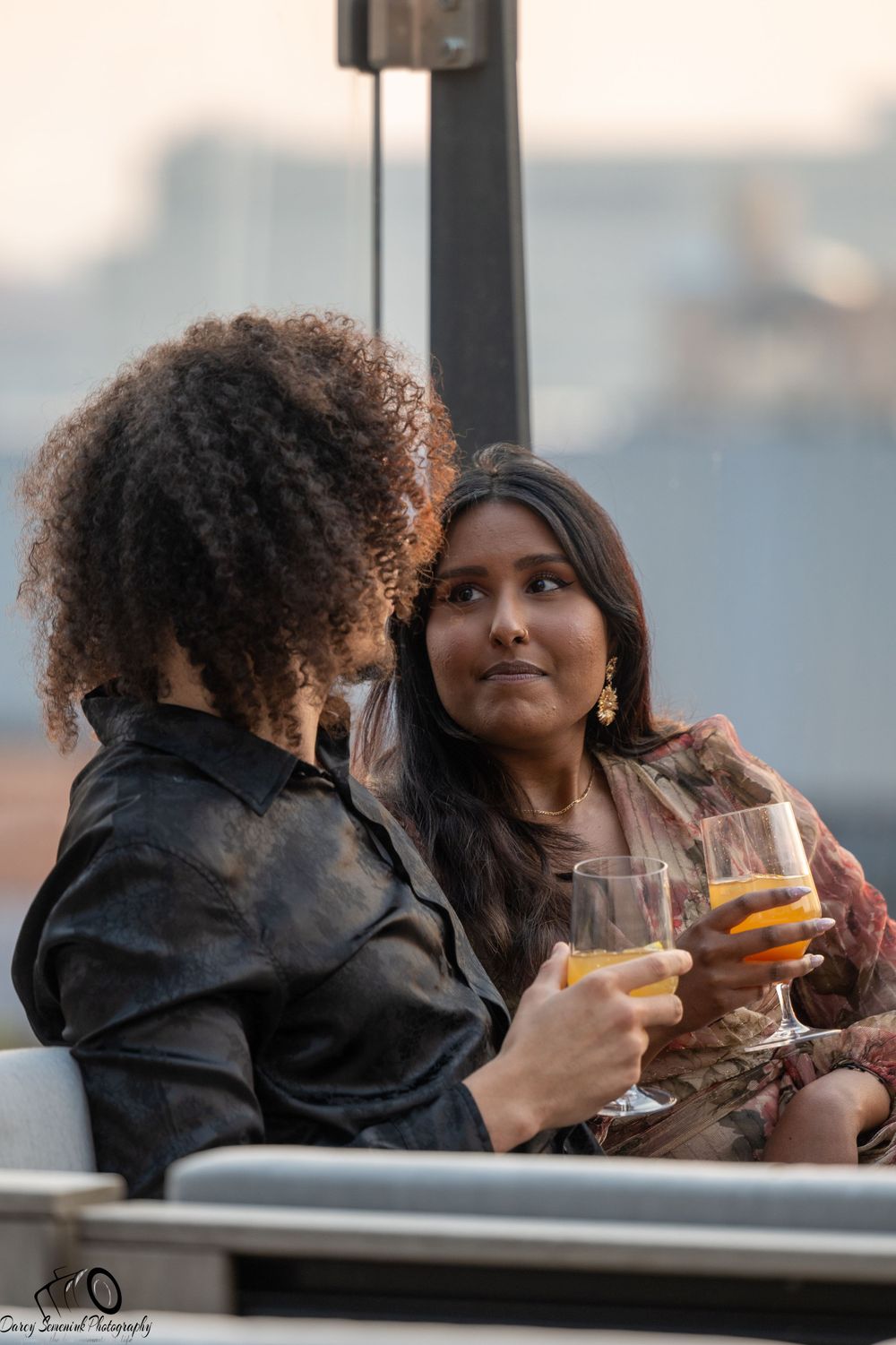 Two friends share drinks and laughter while enjoying a sunset on an outdoor patio.