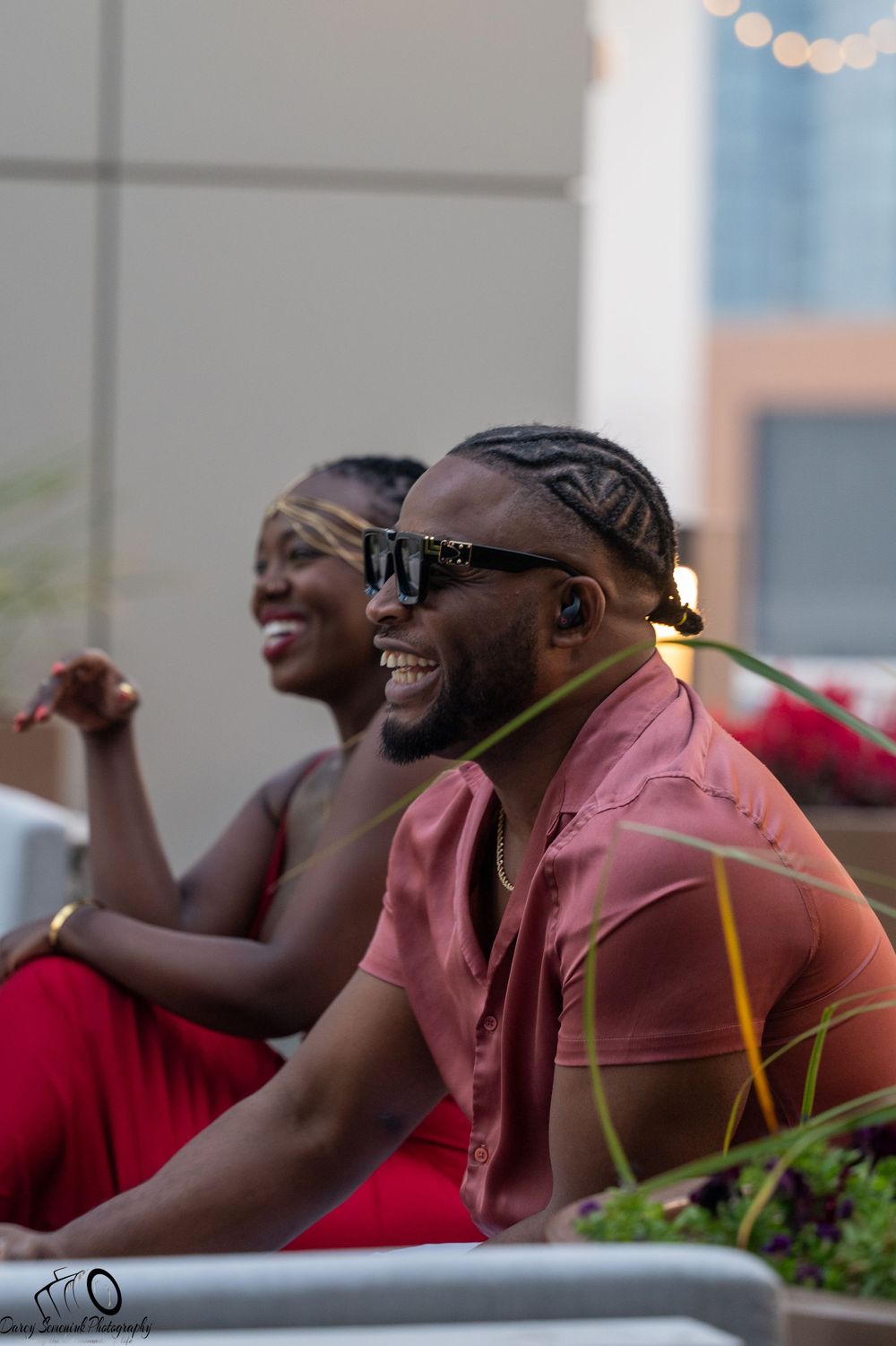 Two people in pink and red shirts laugh together while sitting outdoors.