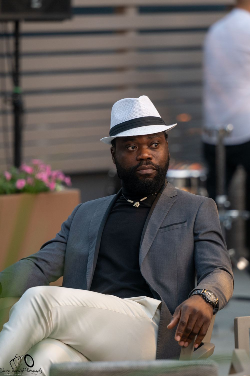 A person in a gray suit and black shirt sits comfortably on a white chair in an outdoor setting.