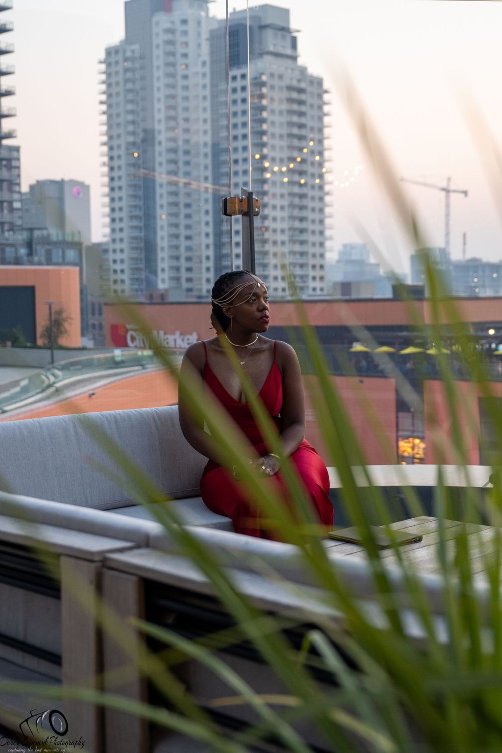 A person in a red dress sits on a rooftop terrace with city high-rises in the background at dusk.