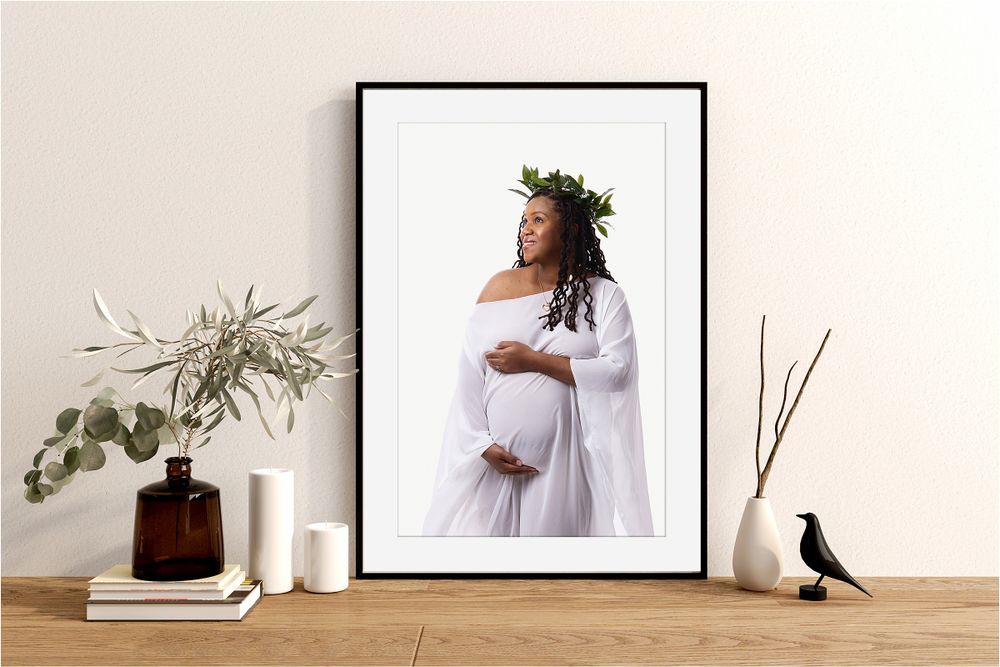 Matted maternity portrait of Black mother in white fabric drape, framed and displayed on tabletop with vases and bird figure